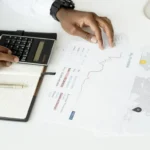 Seseorang menganalisis dokumen keuangan menggunakan kalkulator untuk perencanaan investasi dan perhitungan laba